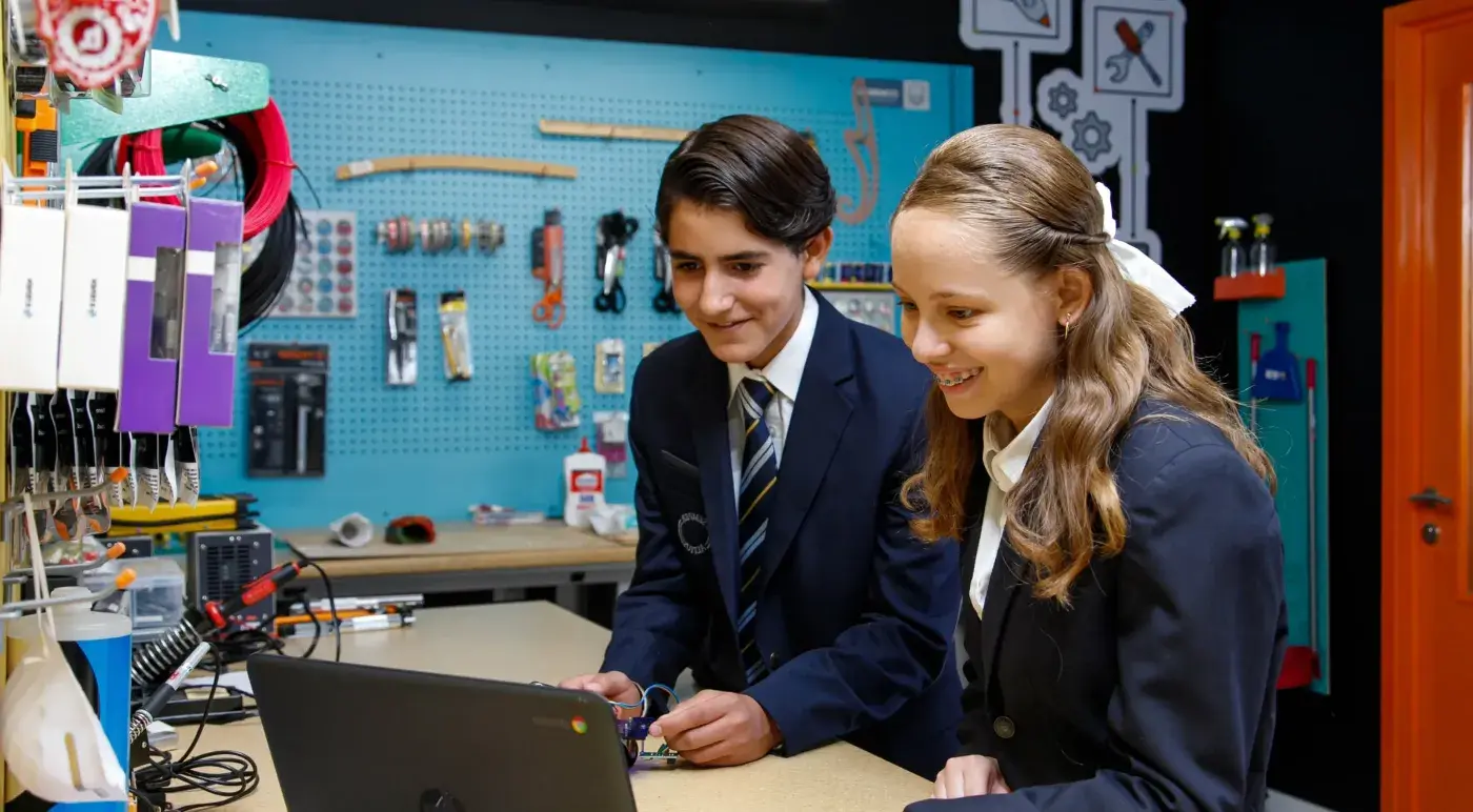 Dos estudiantes de uniforme trabajan juntos en una laptop en un laboratorio de tecnología con herramientas y componentes electrónicos.