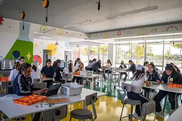 Estudiantes trabajan en una clase de tecnología con laptops y materiales naranjas en aulas modernas y luminosas.