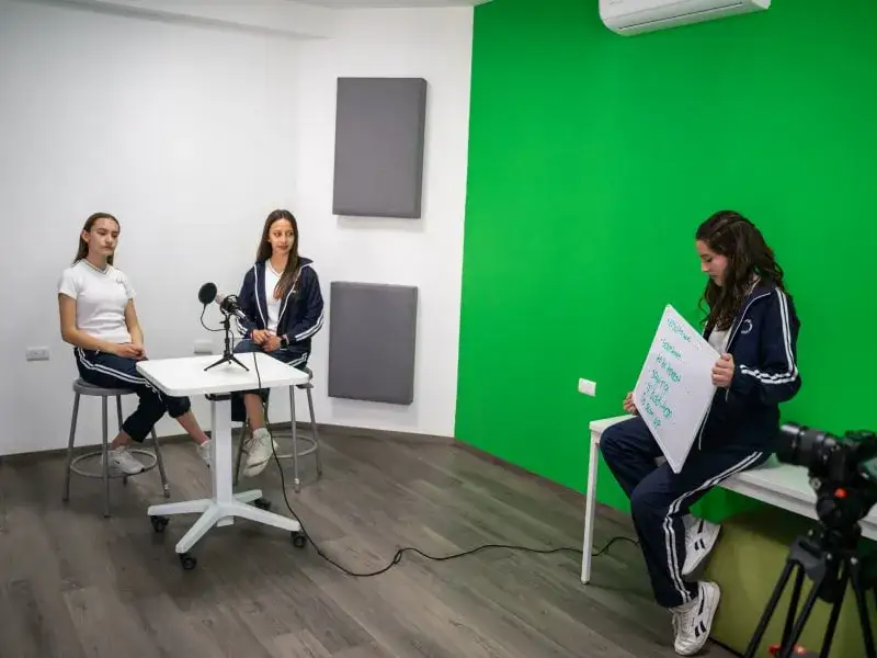 Tres estudiantes en estudio de grabación con pared verde, realizando entrevista con micrófono y cámara